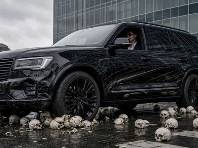 Een dure wagen rijdt over schedels, de bestuurder onbewogen achter zijn zonnebril.